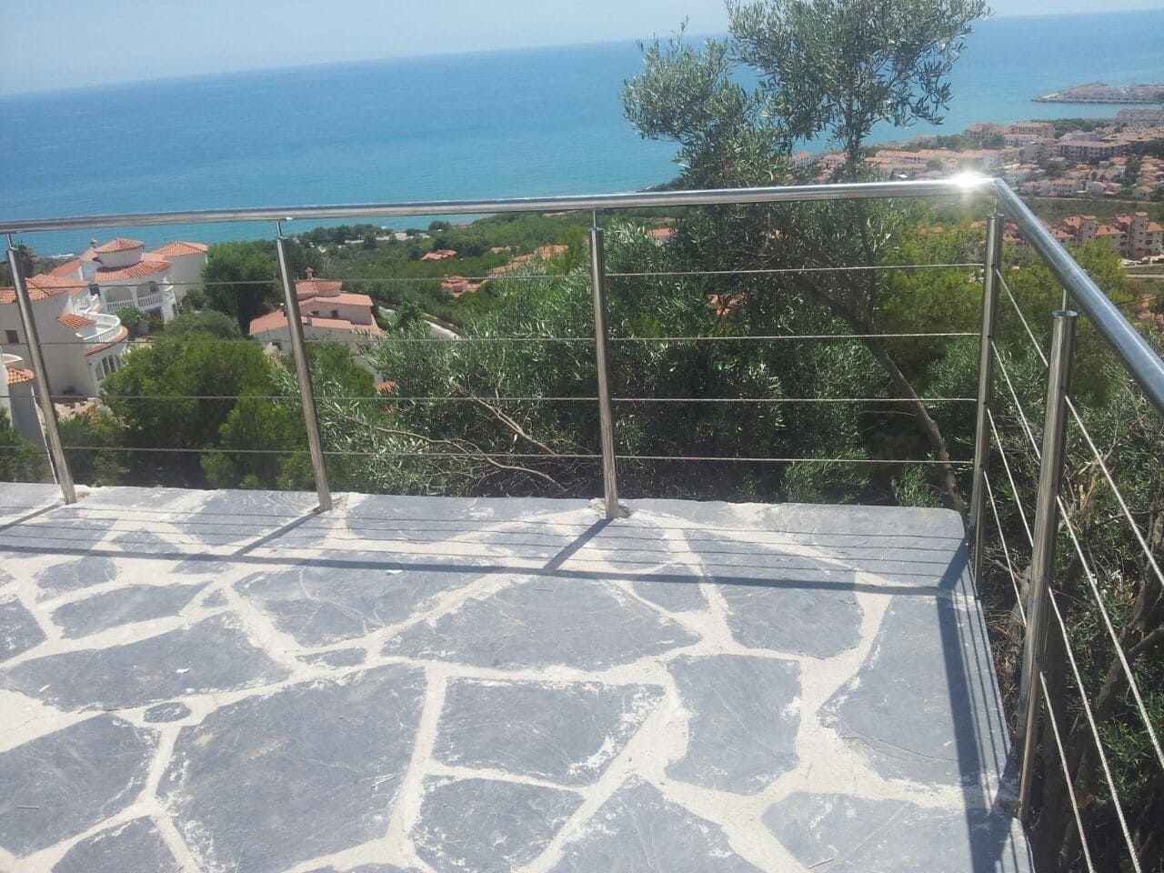 Terraza de piedra con vistas a un pueblo costero y al mar azul, con barandilla de cristal y un árbol en primer plano.