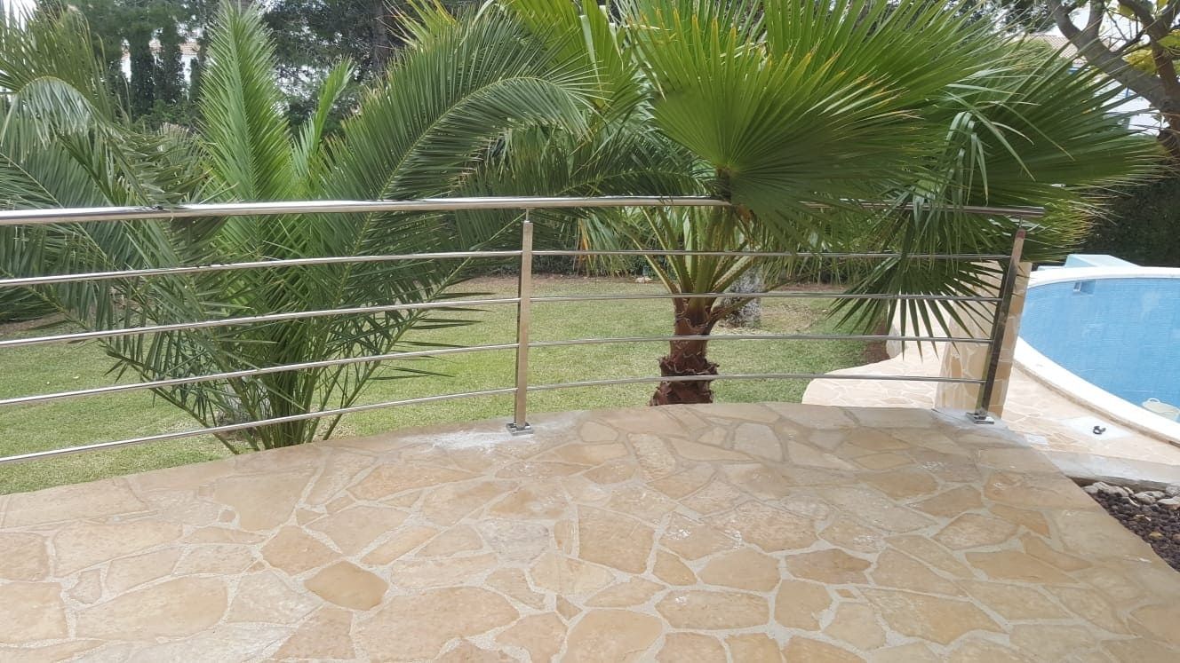 Patio de piedra con barandilla de metal, palmeras y una piscina al fondo.