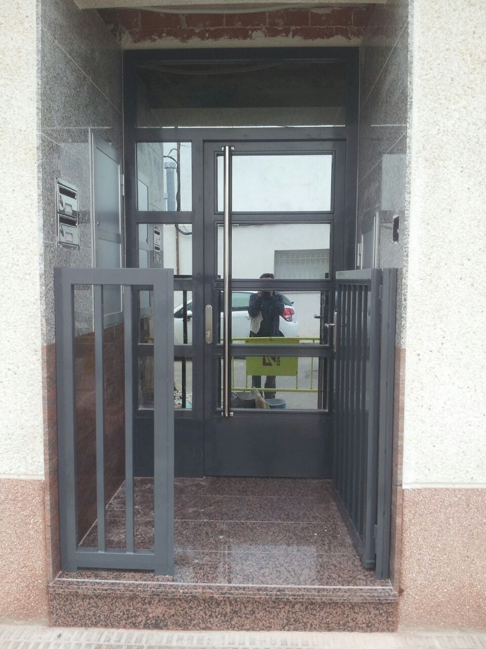 Entrada abierta con verja metálica que da a un vestíbulo interior y puertas de cristal al otro lado.