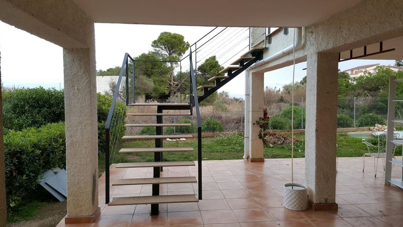 Patio cubierto con escaleras que conducen a un jardín, suelo de baldosas y columnas blancas bajo un cielo nublado.