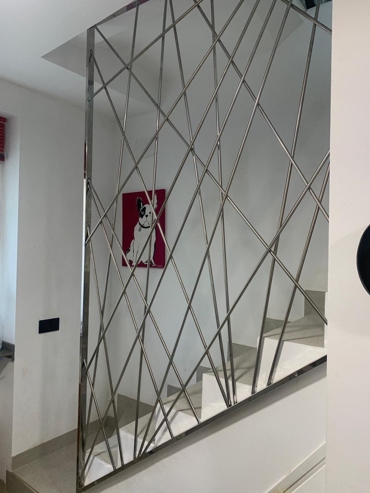 Mampara decorativa de celosía metálica en un pasillo interior blanco con una obra de arte mural roja detrás.