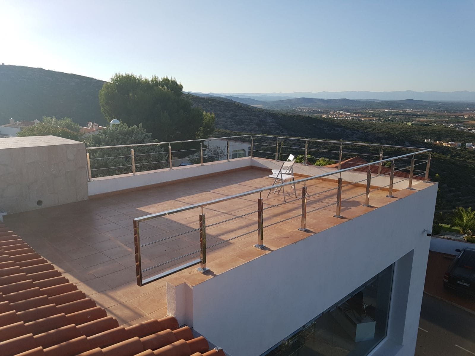 Terraza en la azotea con barandilla de cristal, borde de tejado de tejas rojas y vistas a la ciudad en la ladera al atardecer.