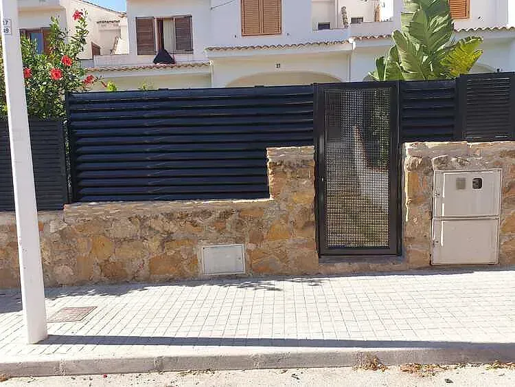 Muro de piedra y puerta negra frente a una casa, con acera de baldosas y caja de servicios públicos de color beige.