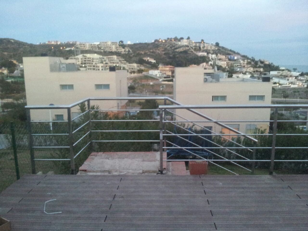 Terraza en la azotea con barandilla metálica con vistas a los edificios y árboles de la ladera al atardecer.