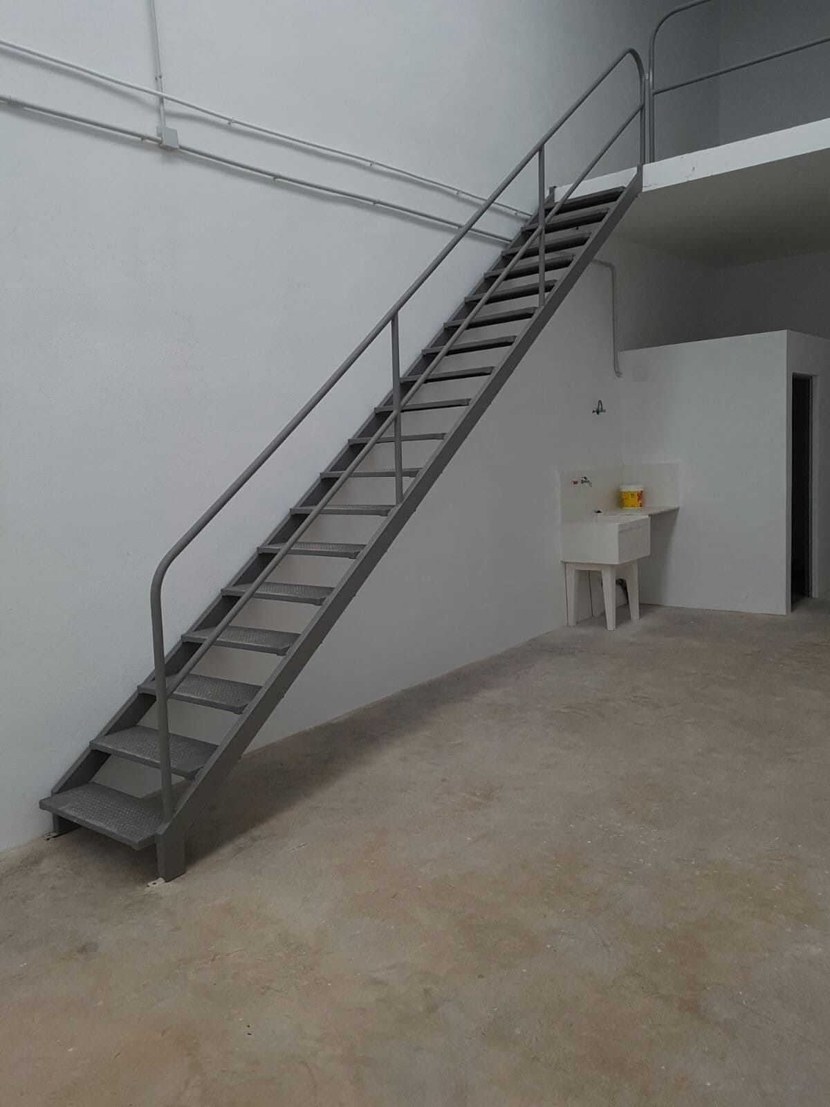 Escalera metálica en una habitación blanca y austera con suelo de hormigón y una pequeña mesa debajo.