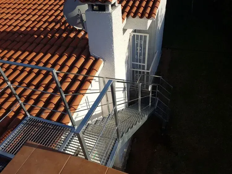 Escalera en la azotea junto a una chimenea blanca, con baldosas de terracota y una pared oscura y sombreada.