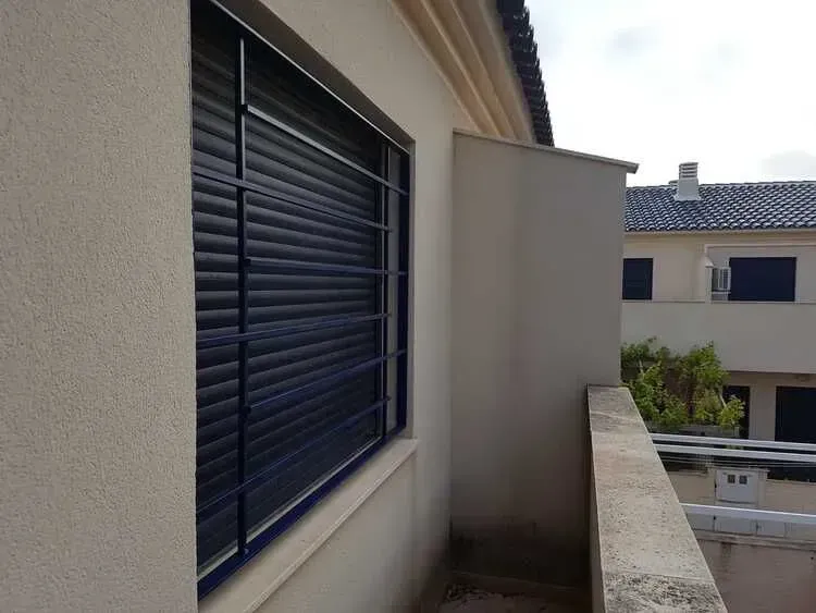 Balcón de apartamento con paredes beige, una ventana estrecha con contraventanas oscuras y vistas a los edificios vecinos.