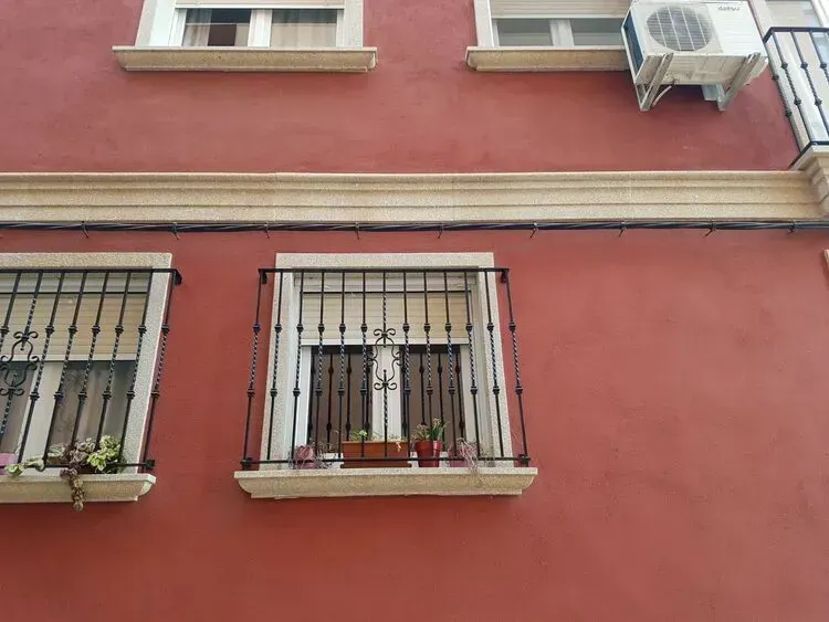 Fachada roja del edificio con ventanas enrejadas, jardineras y una unidad de aire acondicionado.