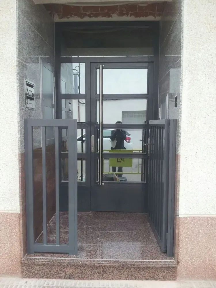 Entrada con verjas metálicas abiertas y una persona reflejada en las puertas de cristal del interior.