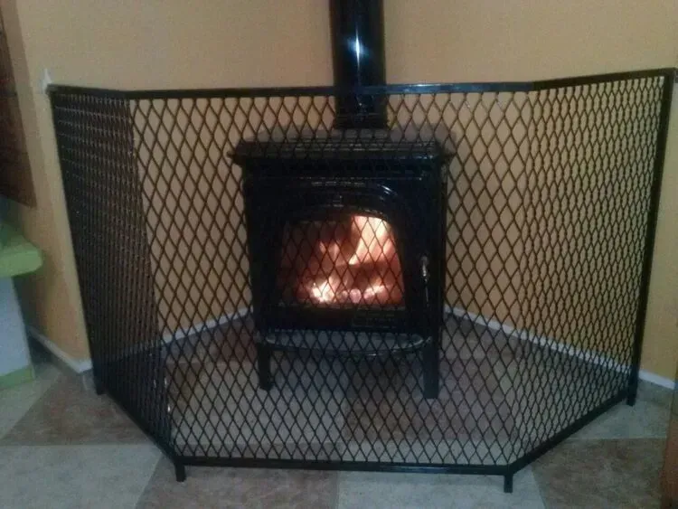 Estufa de leña negra encendida detrás de una pantalla ignífuga curva de malla negra sobre un suelo de baldosas.