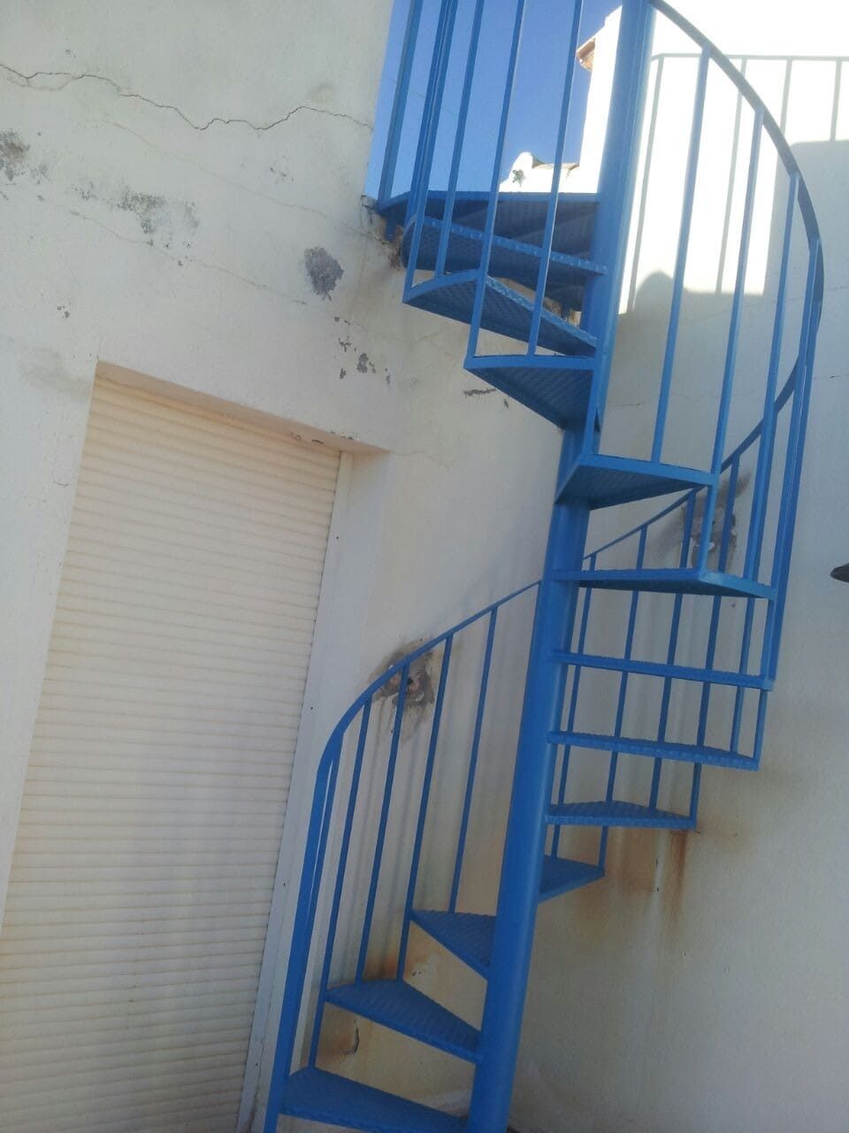 Escalera de caracol de metal azul sobre una pared exterior blanca, junto a una puerta con contraventana cerrada.
