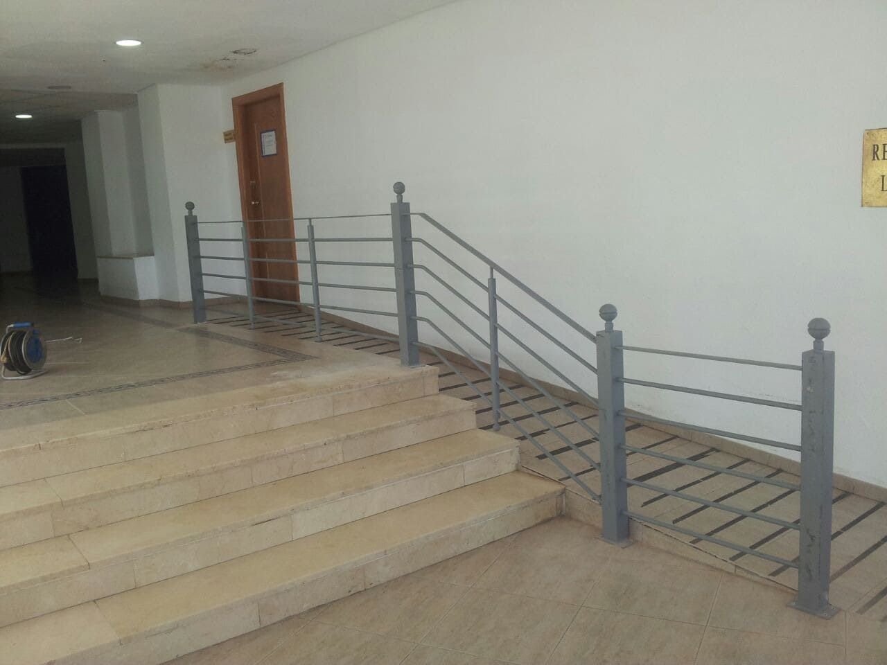 Escalera interior con barandilla gris, suelo de baldosas y una puerta de madera en el pasillo.