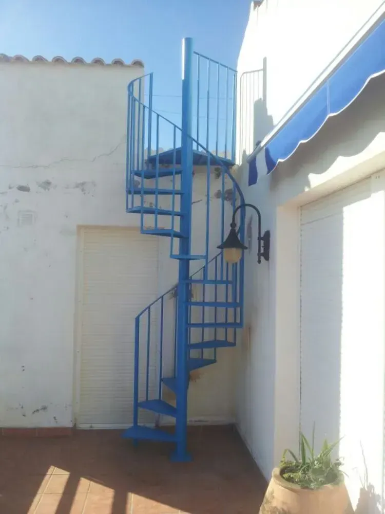 Escalera de caracol azul junto a un edificio de estuco blanco con toldo azul en un patio soleado.