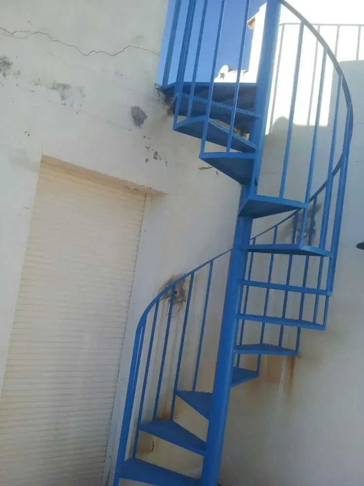Escalera de caracol azul junto a una pared beige y una puerta enrollable blanca en el exterior.