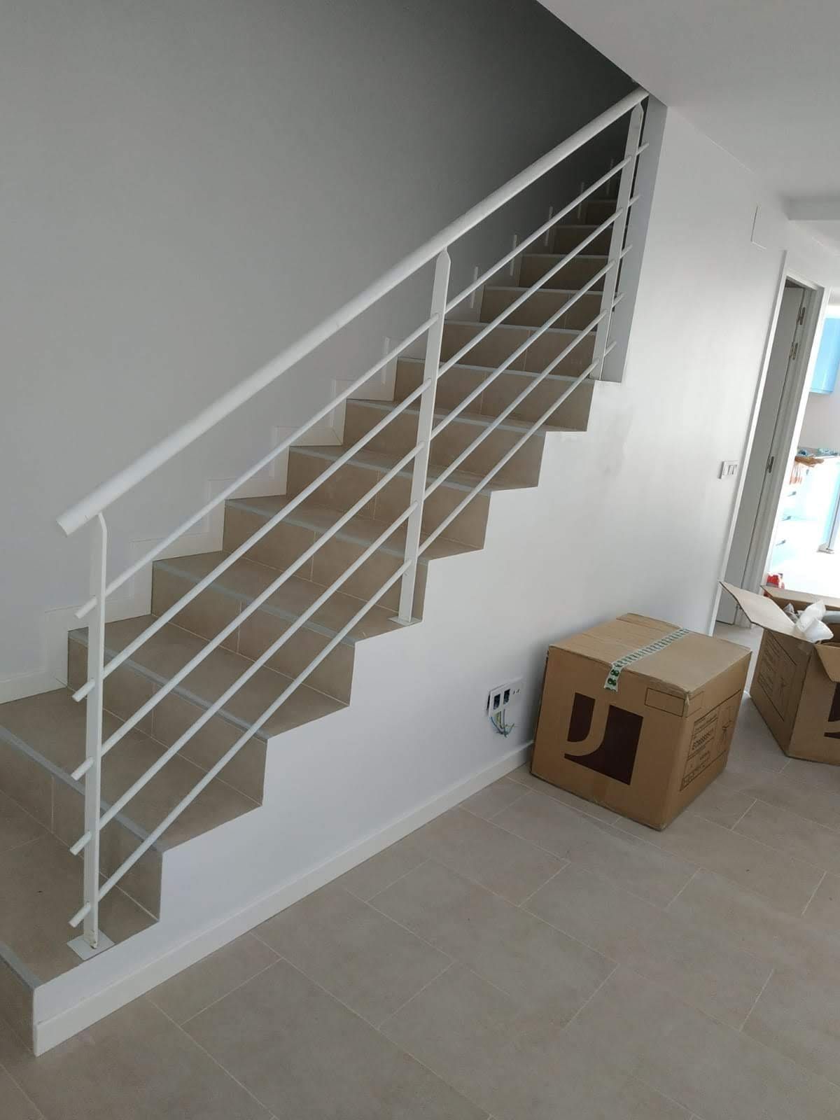 Escalera blanca con barandilla de metal en una habitación luminosa junto a cajas de cartón y suelo de baldosas.
