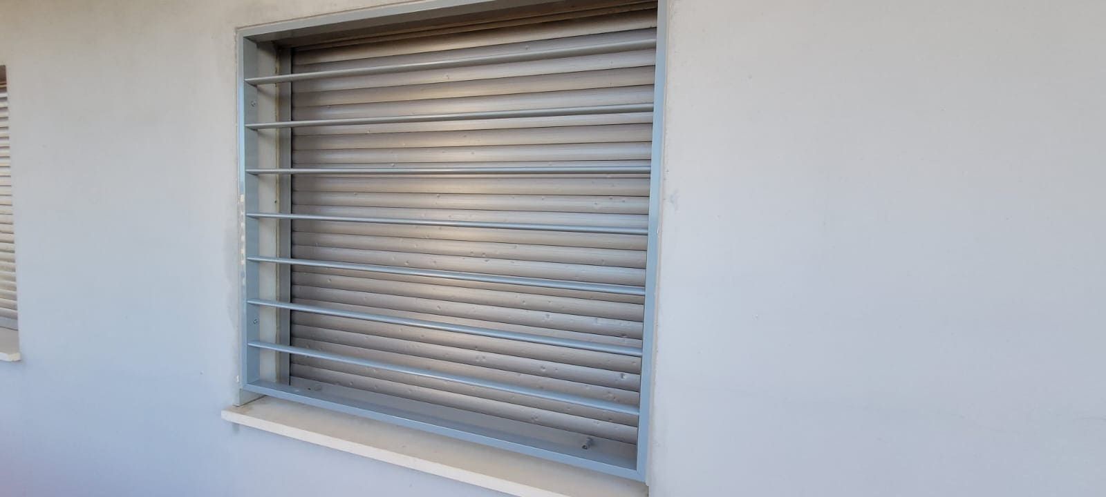 Ventana con persianas metálicas plateadas cerradas, enmarcada en una pared exterior blanca.