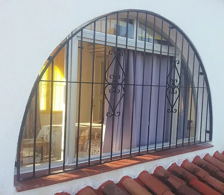 Ventana arqueada con barrotes de metal negro y luz interior cálida sobre tejas rojas.