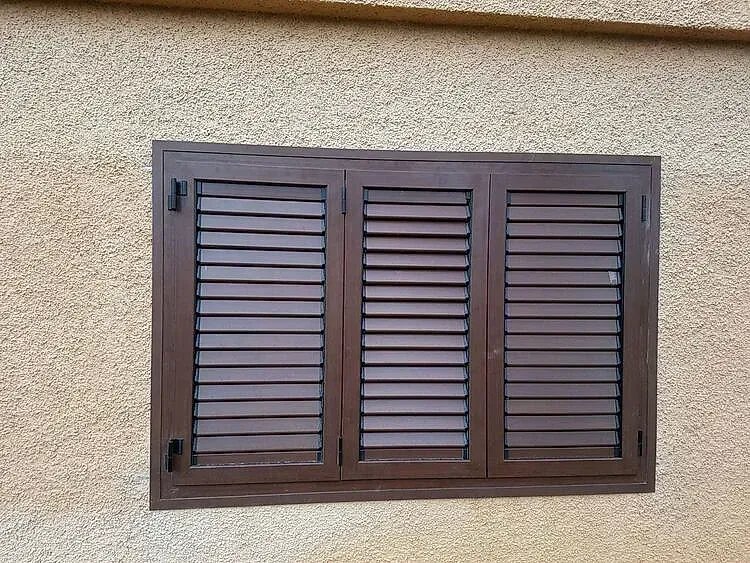 Ventana cerrada con contraventana de madera marrón sobre una pared de estuco beige.