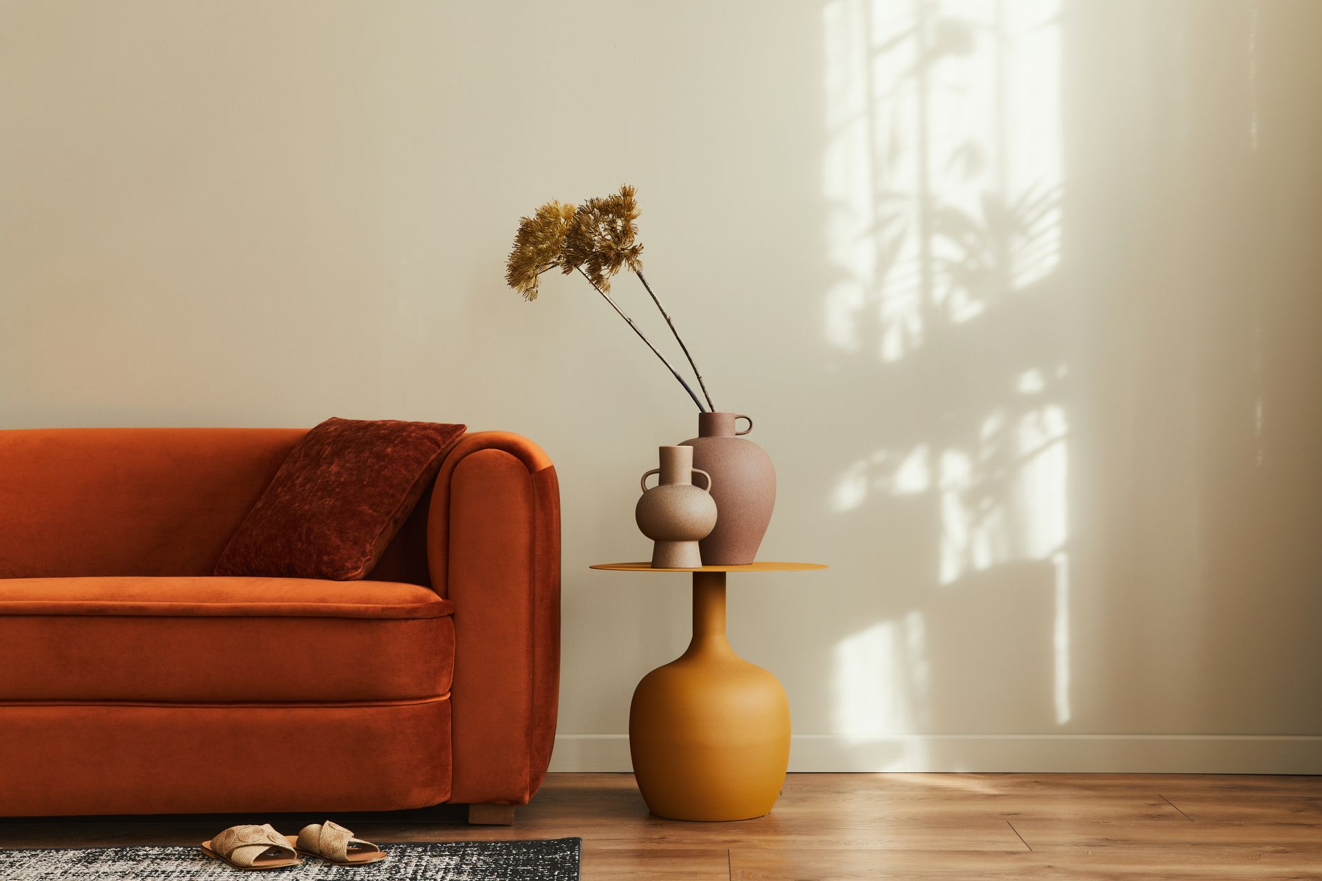 Un canapé en velours orange est placé à côté d'une table d'appoint jaune moutarde avec des vases contenant des fleurs séchées, contre un mur crème.