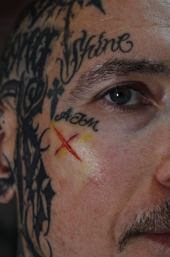 Rostro de un hombre con tatuajes cerca del ojo y la sien, incluyendo una marca roja en forma de X debajo del ojo.