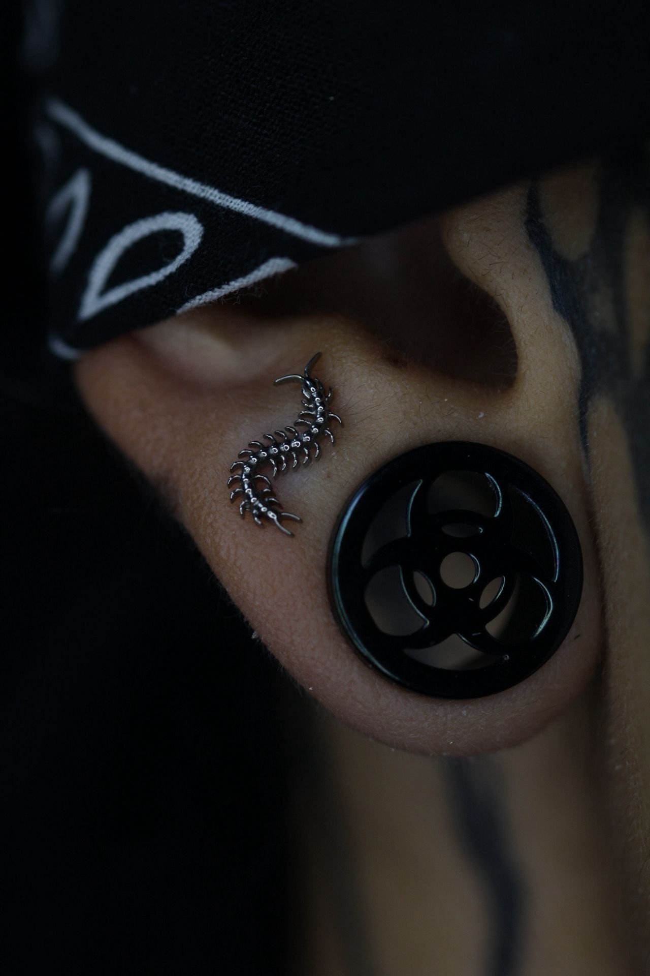 Primer plano de un pendiente de tapón negro con un dije de serpiente plateada y pintura facial oscura sobre la piel.