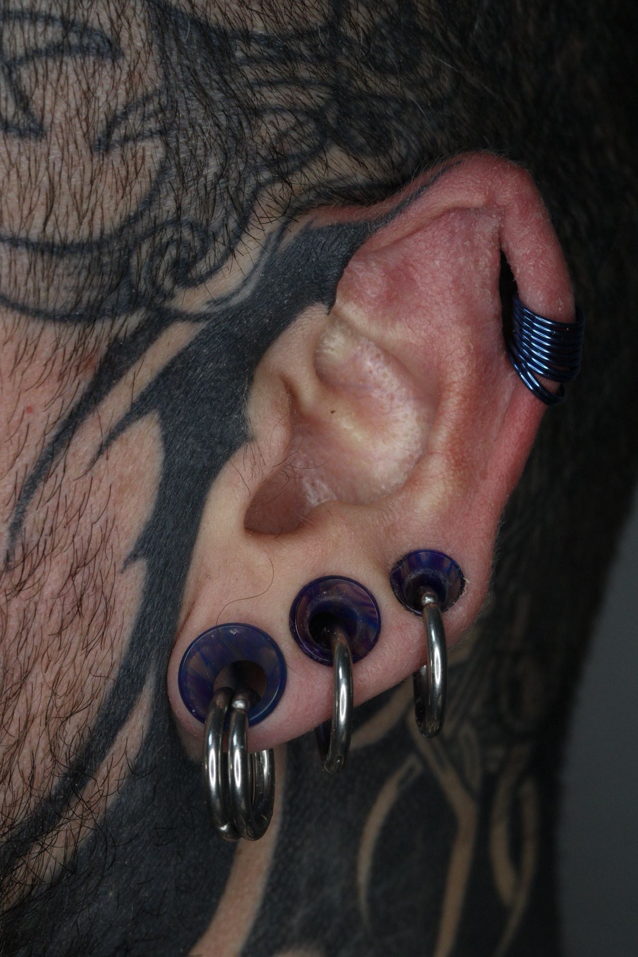 Primer plano de una oreja con múltiples perforaciones negras y plateadas y tatuajes cercanos.