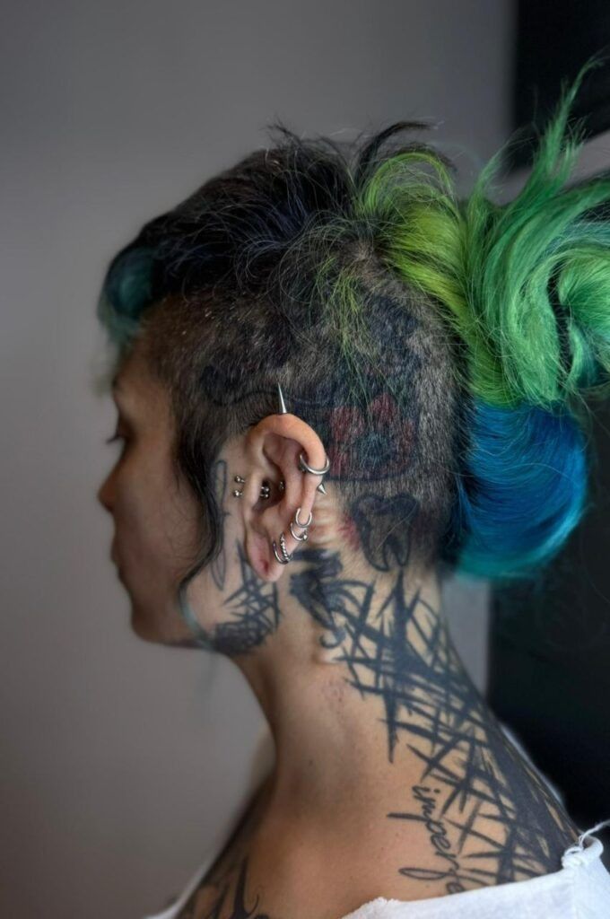 Perfil lateral de una persona con cabello verde y azul y tatuajes en el cuello sobre un fondo gris.