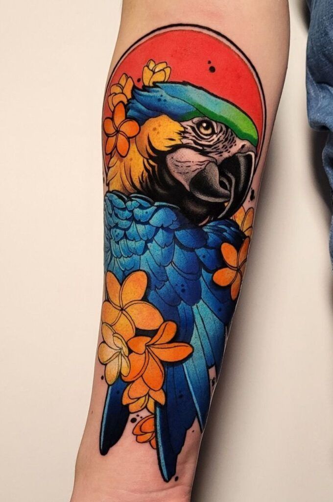 Tatuaje de un loro colorido en la piel con plumas azules, flores naranjas y un fondo rojo.