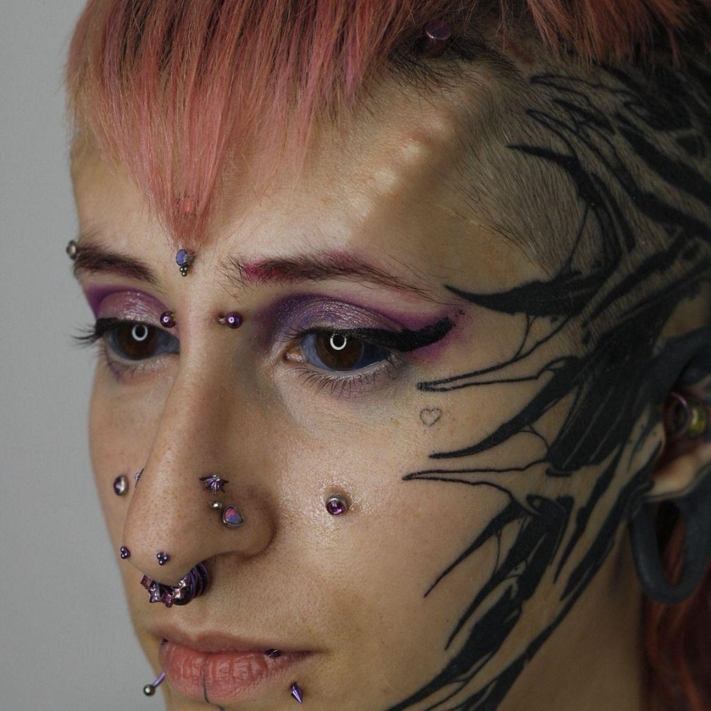 Persona con cabello rosa, maquillaje de ojos morado, piercings faciales y pintura facial negra sobre un fondo liso.