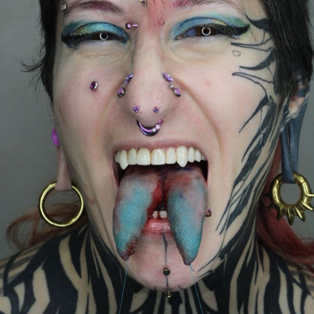Primer plano de un rostro con muchos piercings, lengua azul afuera, pintura facial negra y una expresión feroz.