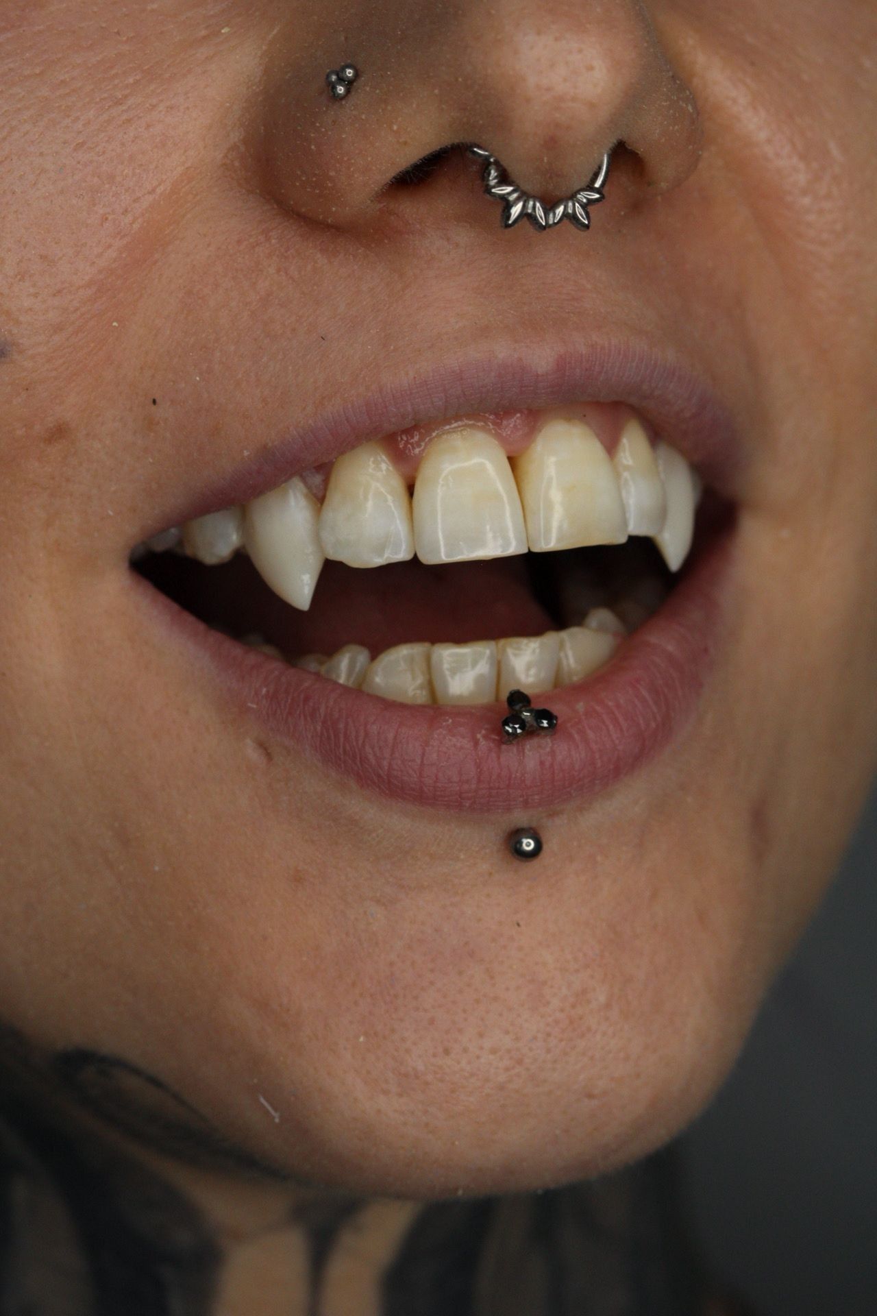 Primer plano de una boca sonriente con piercings plateados y caninos afilados y puntiagudos.