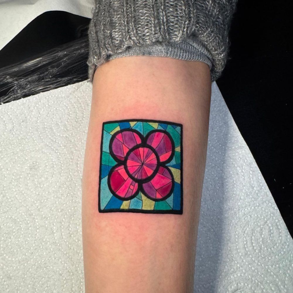 Tatuaje floral colorido en un antebrazo, con pétalos rosas y un borde verde azulado.