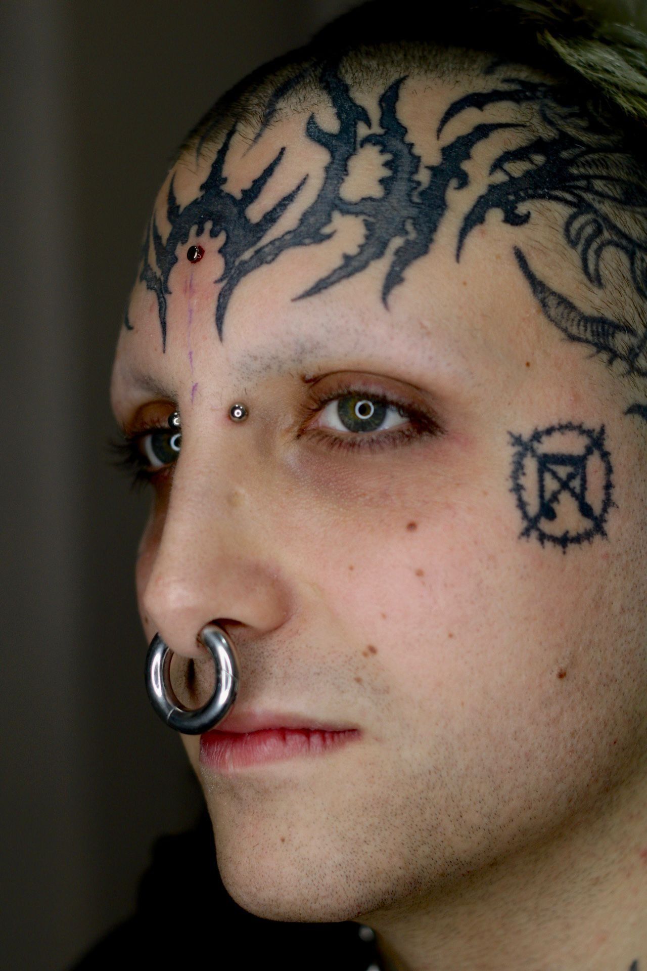 Primer plano del perfil de una persona con tatuajes faciales y un piercing en la nariz, mirando hacia la izquierda.