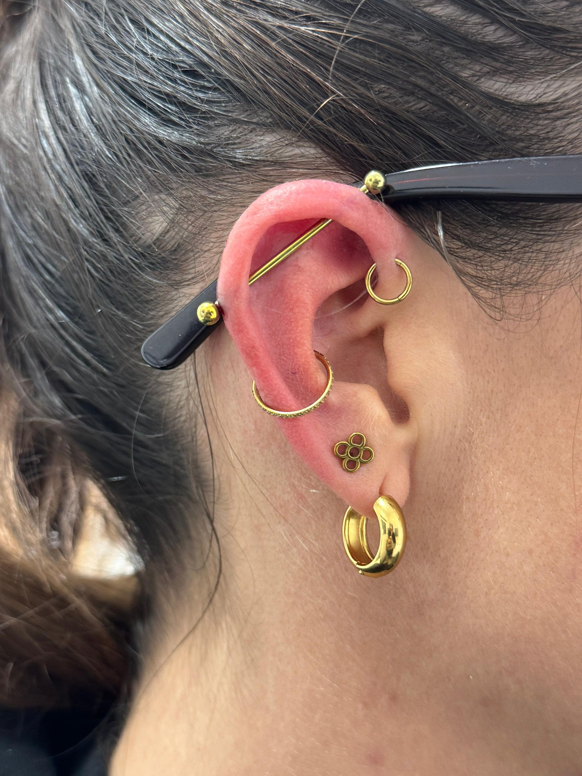 Primer plano de una oreja con varios pendientes de oro y una pinza negra para el cabello que sujeta el cabello oscuro.
