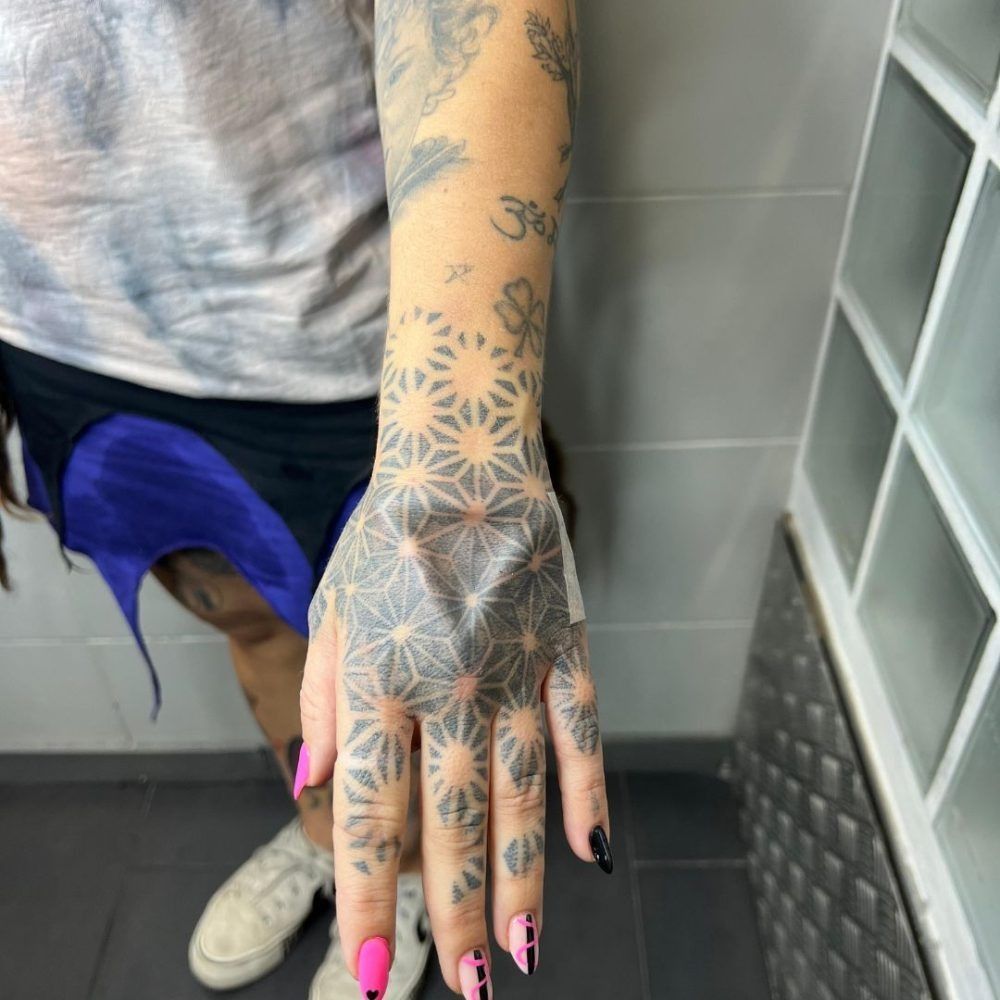Mano tatuada con uñas negras y rosas, apoyada sobre el suelo de baldosas de un baño, cerca de un lavabo y un espejo.