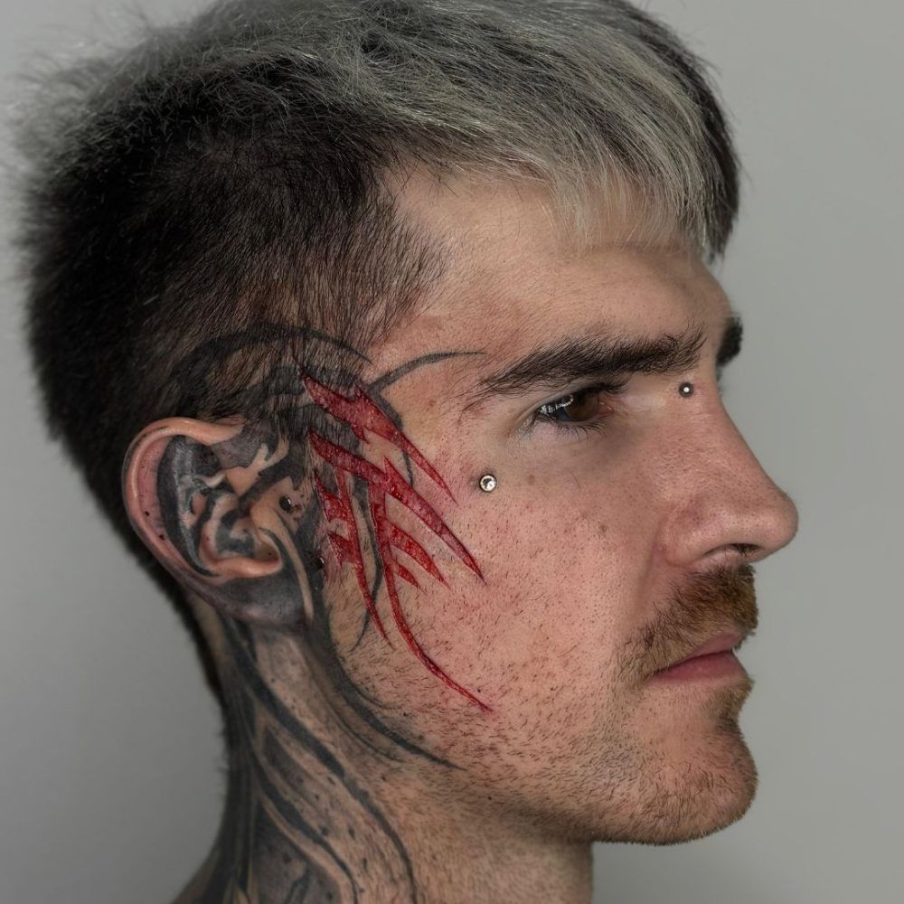Perfil de un hombre con cabello gris y tatuajes faciales, mirando hacia la derecha.