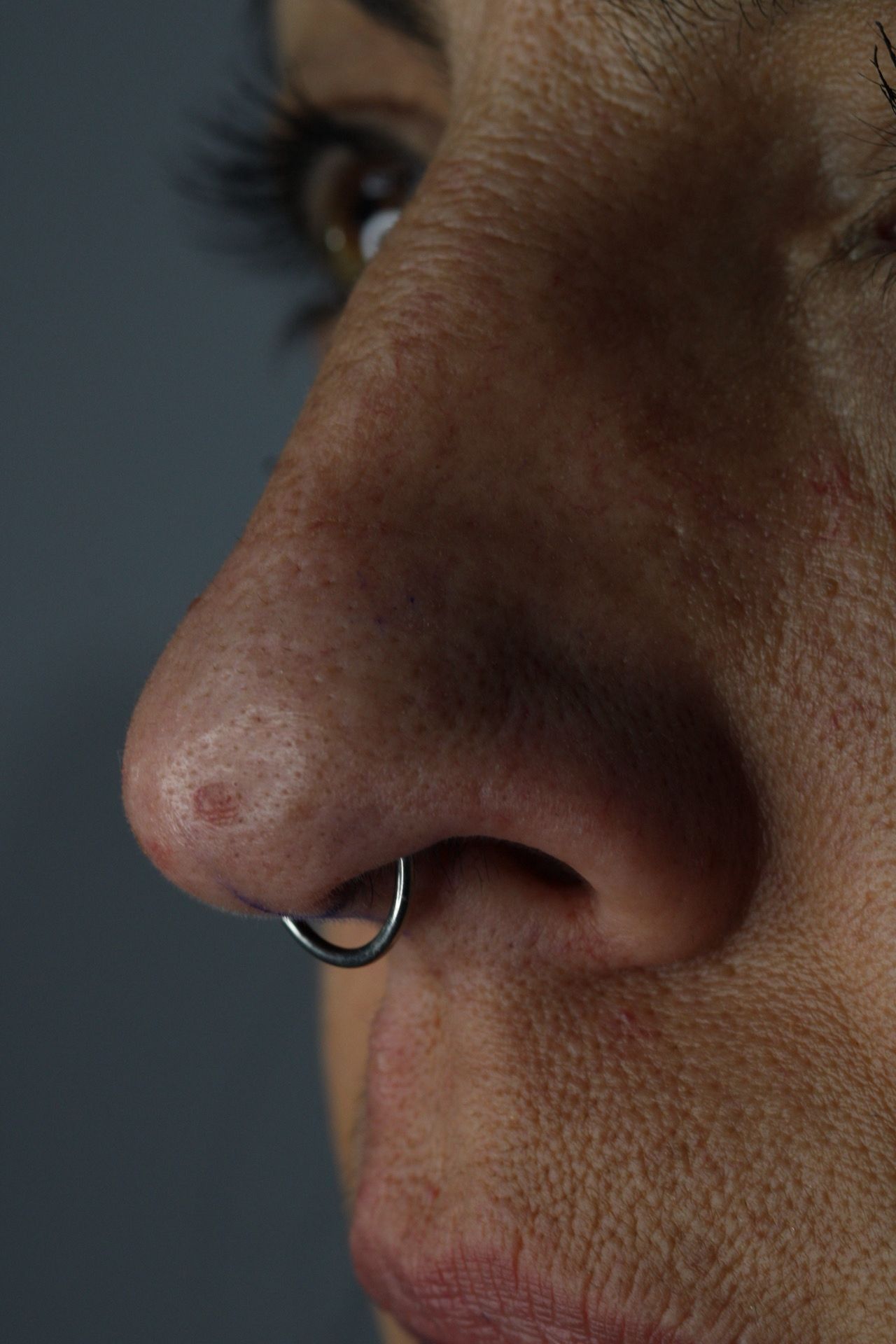 Primer plano de la nariz de una persona con un aro nasal plateado sobre un fondo oscuro.
