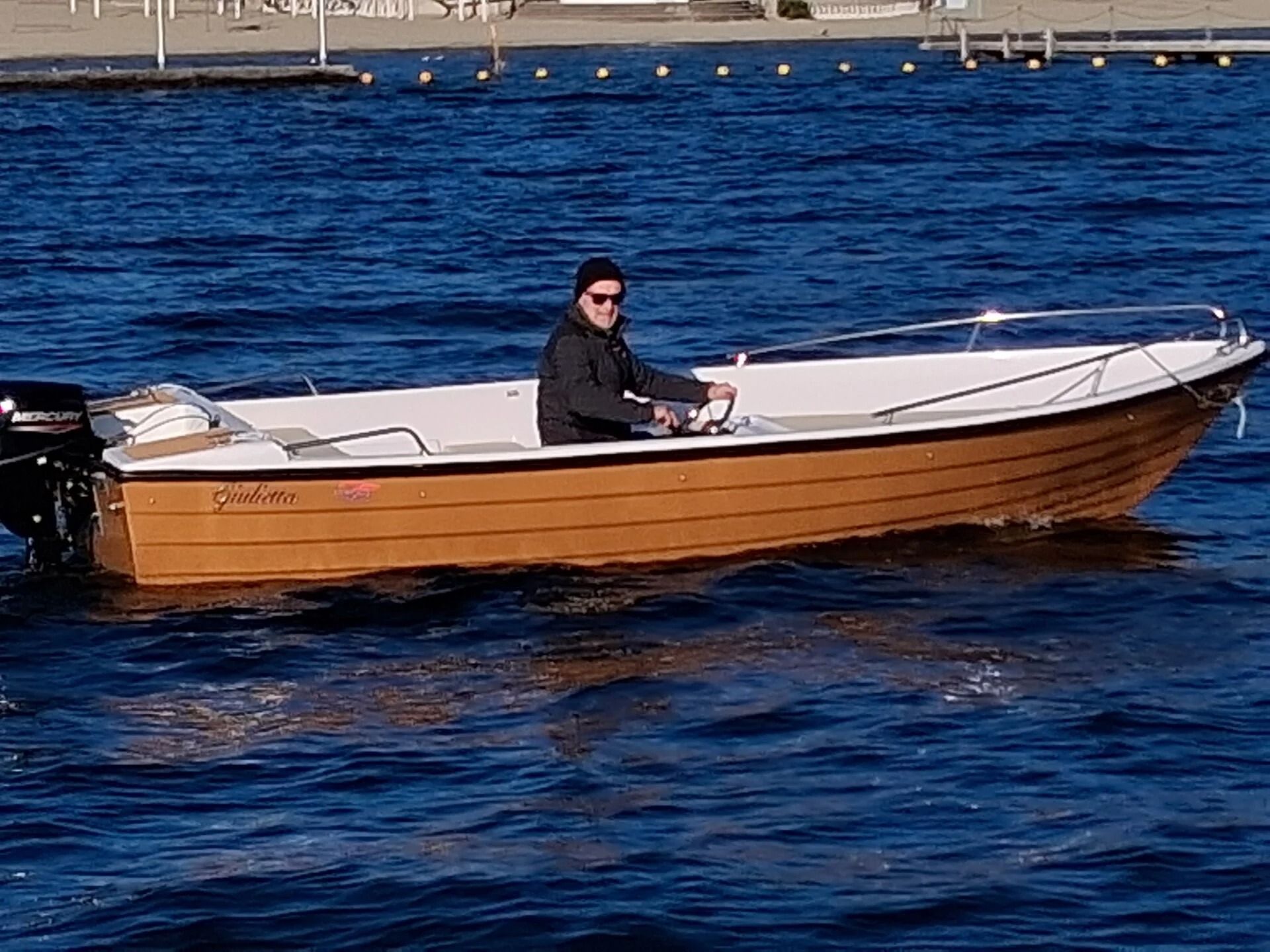 Ti Yachts Sagl 
Una persona guida una piccola barca a motore in legno su acque azzurre, con un porto sullo sfondo.