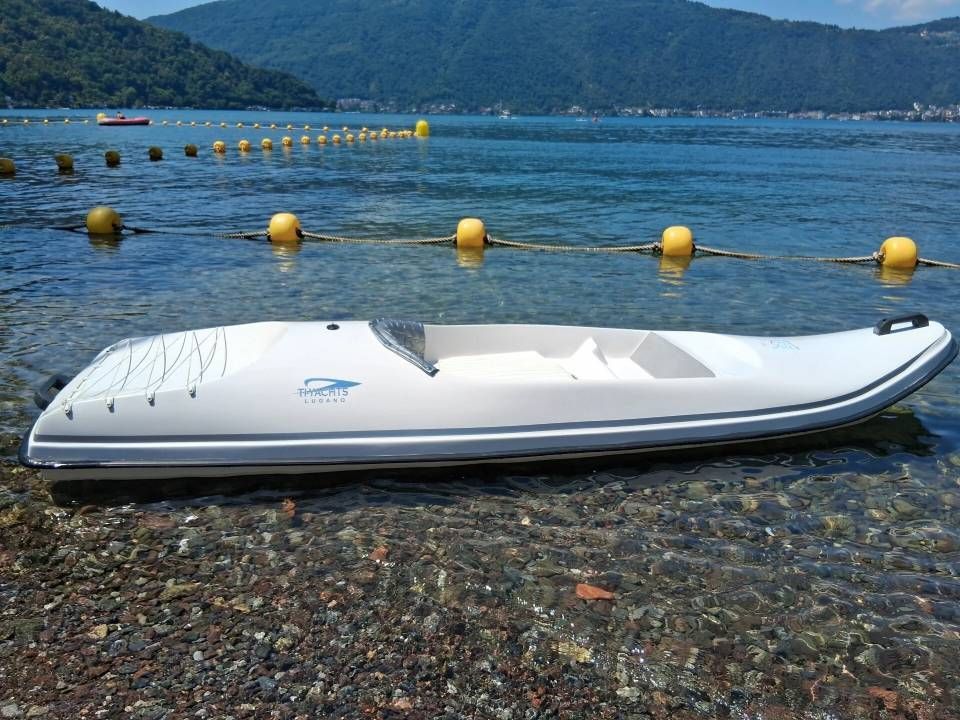 Un gommone bianco su una spiaggia di ciottoli, accanto a acque cristalline di un blu intenso, con una boa gialla e montagne sullo sfondo.