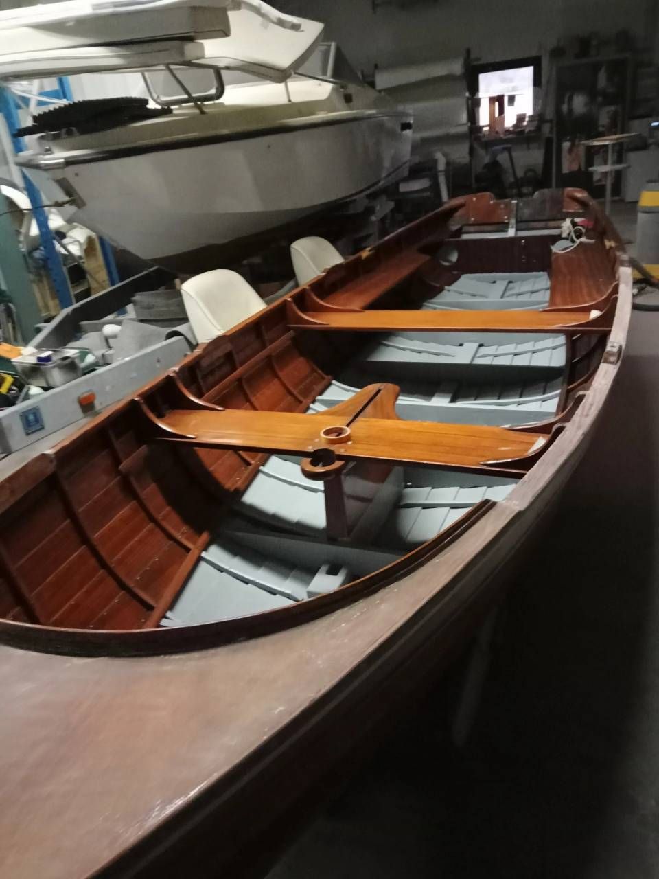 Barca a remi in legno con sedili arancioni all'interno di un'officina accanto a una barca più grande.