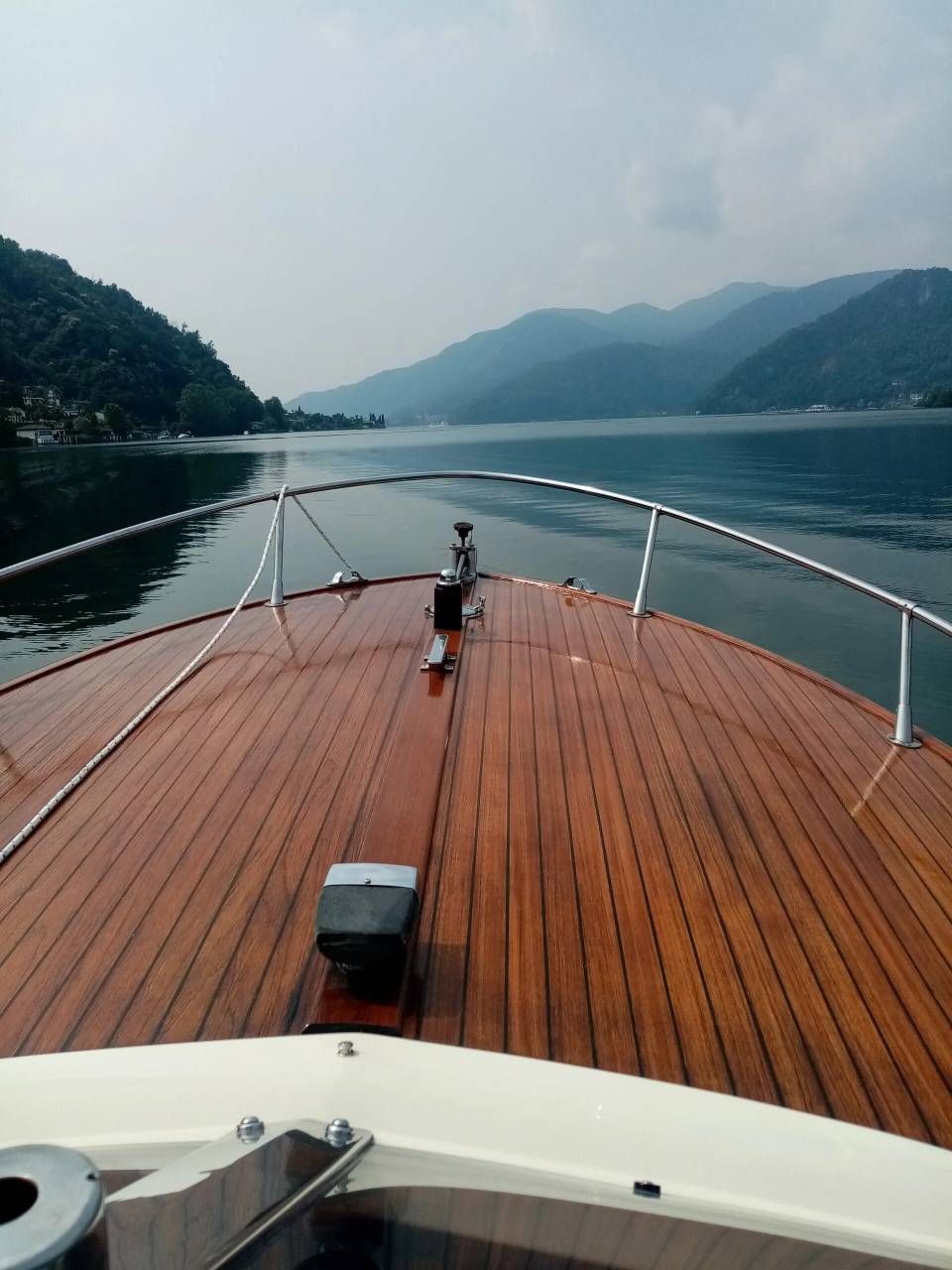 Vista dal ponte di una barca su un tranquillo lago di montagna, con assi di legno e colline nebbiose all'orizzonte.