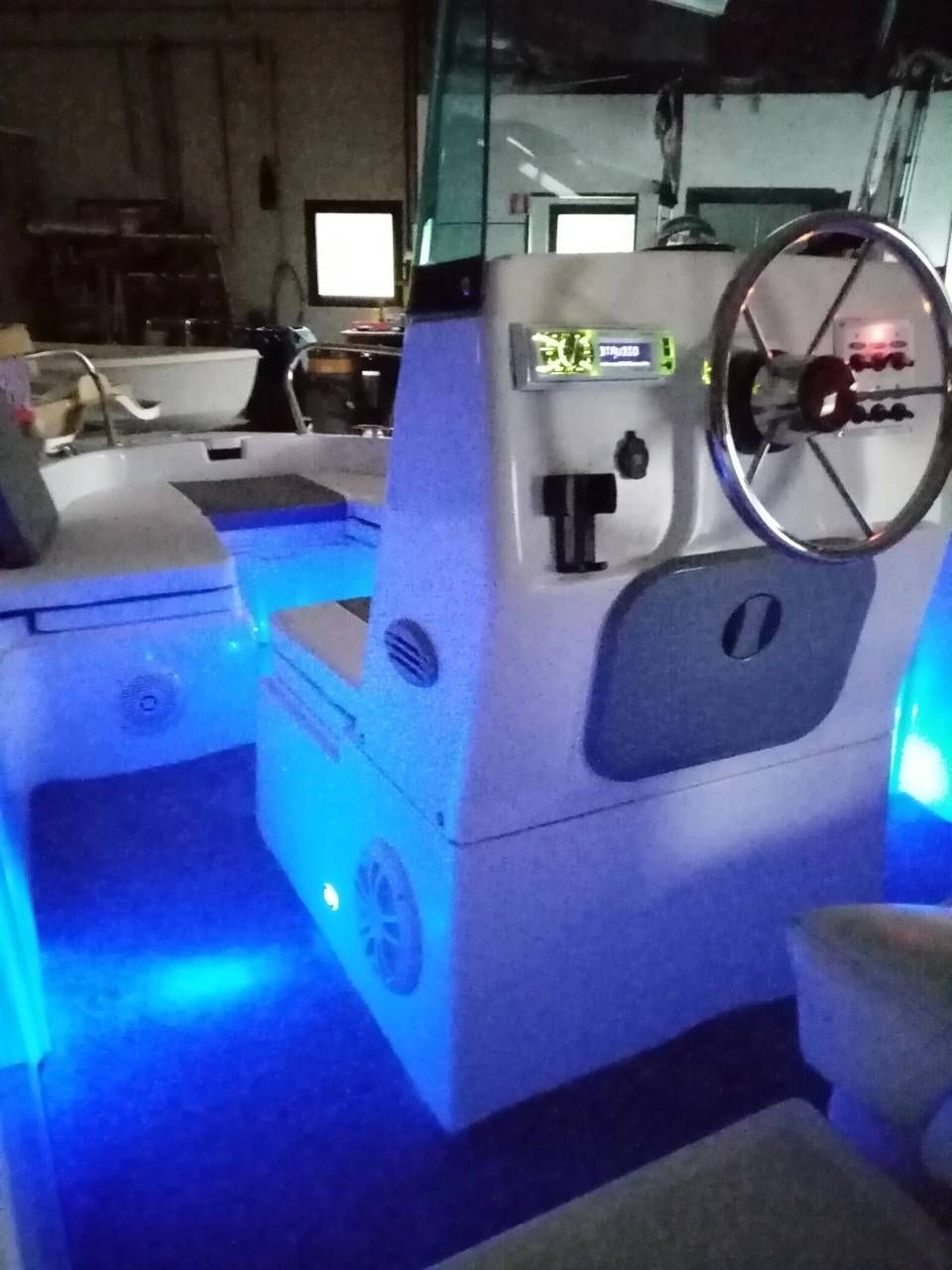 La postazione di comando di una piccola imbarcazione, con volante e cruscotto illuminati di blu in una cabina poco illuminata.