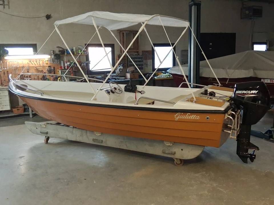 Piccola barca a motore in legno con tettuccio bianco, parcheggiata al coperto su un cavalletto con ruote.