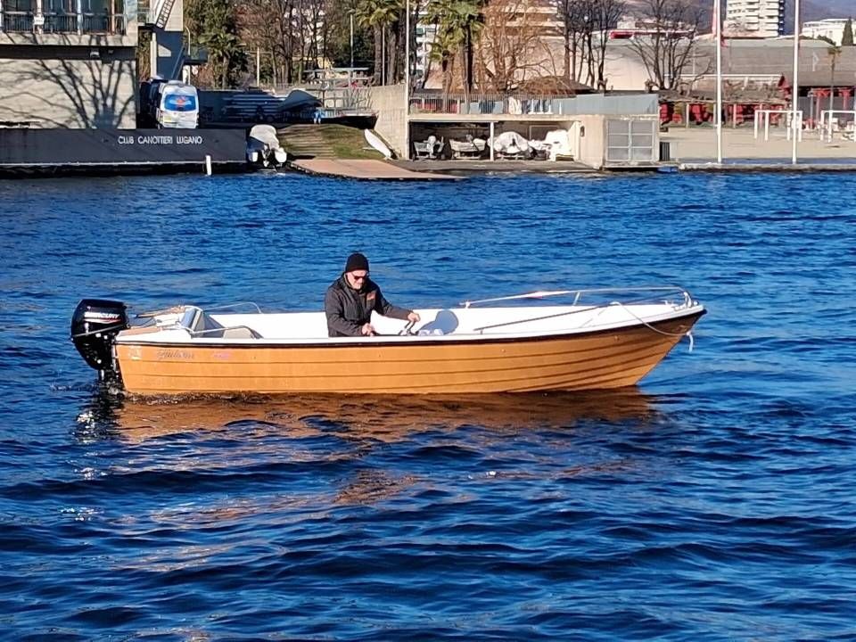 Piccola imbarcazione a motore in legno su acque azzurre con una persona seduta all'interno vicino a un molo sul lungomare.