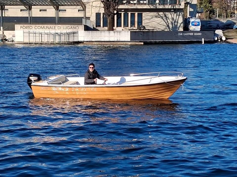 Piccola imbarcazione a motore in legno su acque azzurre vicino a un edificio sul lungomare, con una persona seduta a bordo.