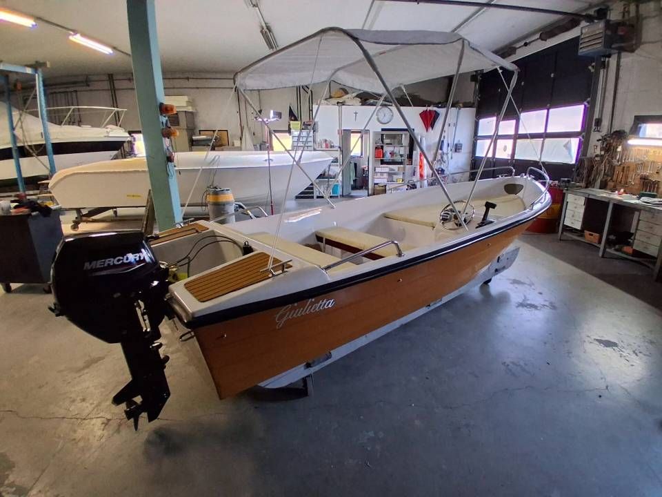 Piccola barca a vela con scafo color sabbia in un'officina, con motore fuoribordo e sartiame sopra la testa.