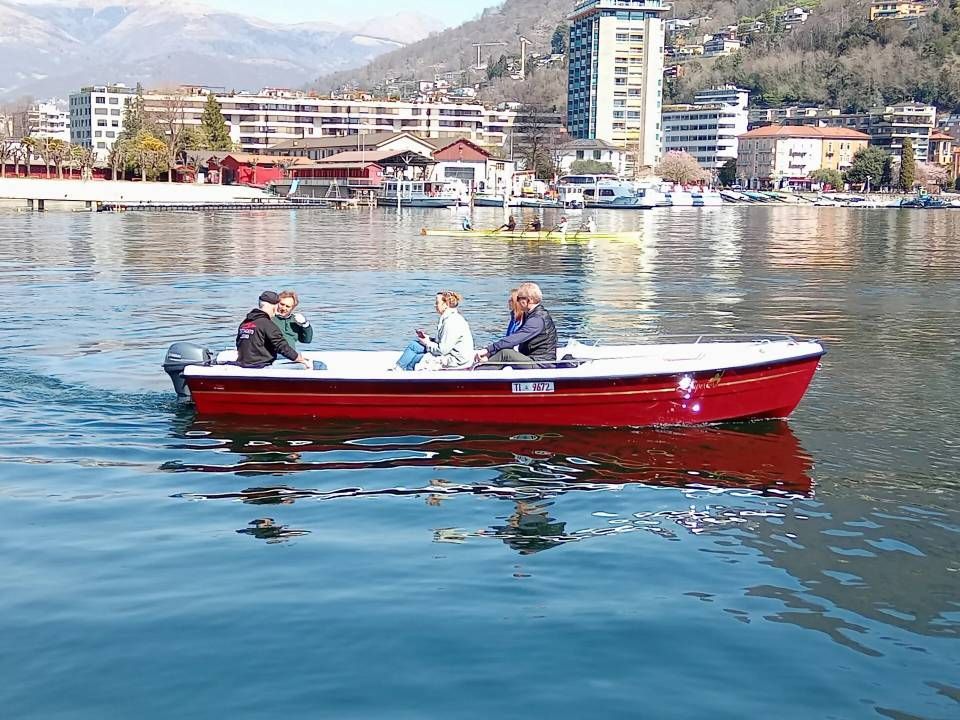 Tre persone viaggiano su un motoscafo rosso su un lago tranquillo, con una città e le montagne sullo sfondo.