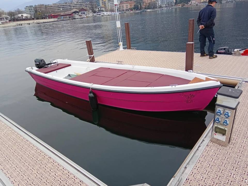 Una piccola imbarcazione magenta e bianca è ormeggiata a un molo su acque calme, con una persona in piedi nelle vicinanze.