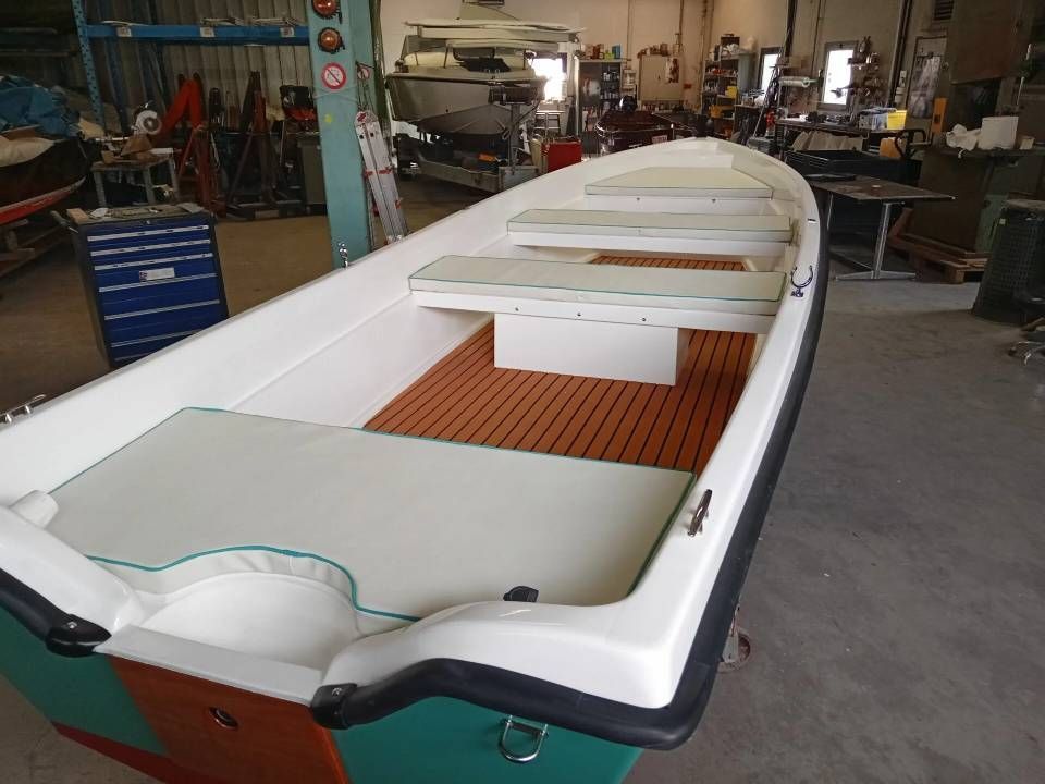 Scafo di barca bianco in costruzione in un'officina, con ponte in legno e panche all'interno.