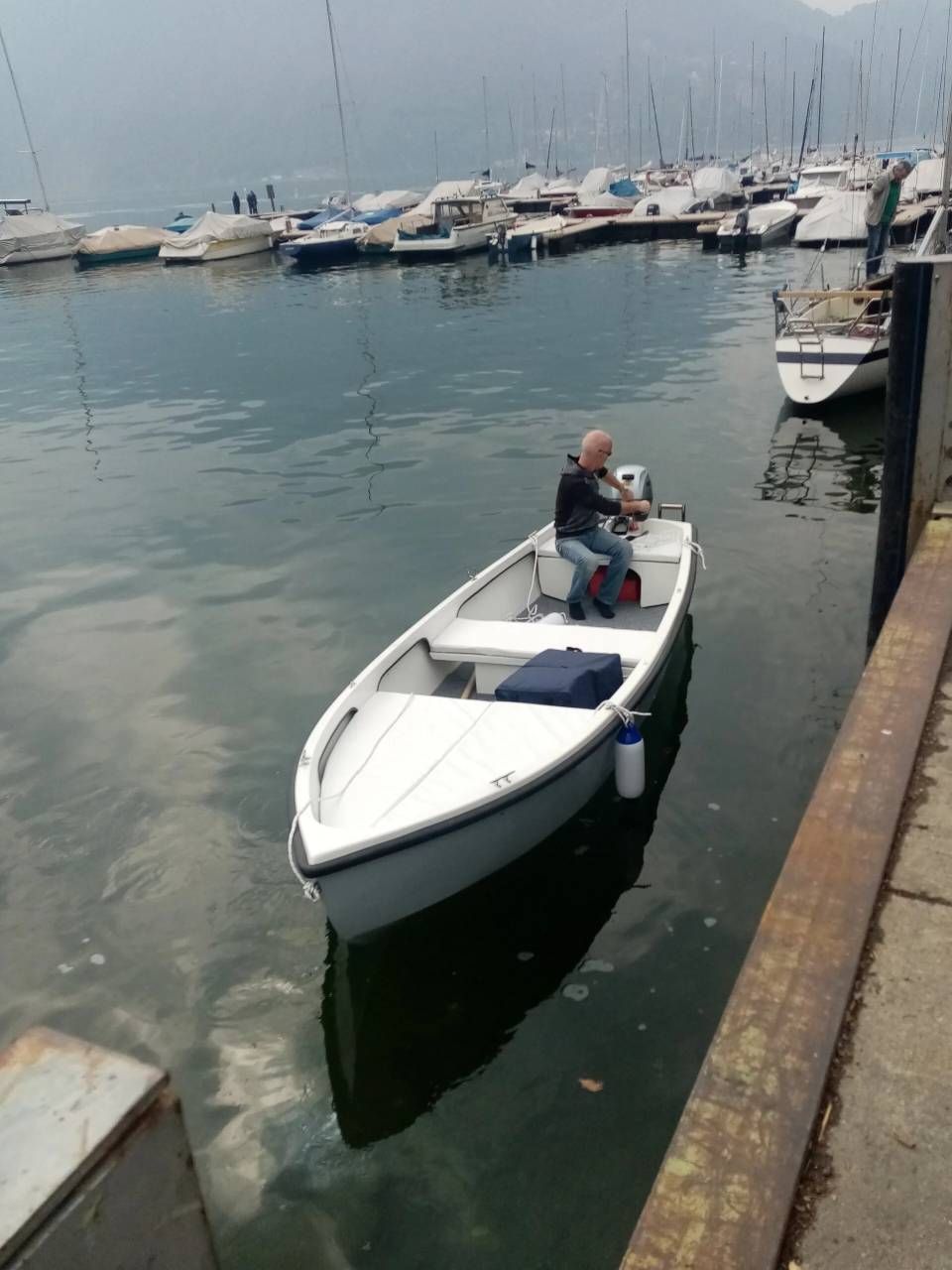 Piccola imbarcazione a motore bianca con una persona seduta in un porticciolo accanto a yacht ormeggiati.